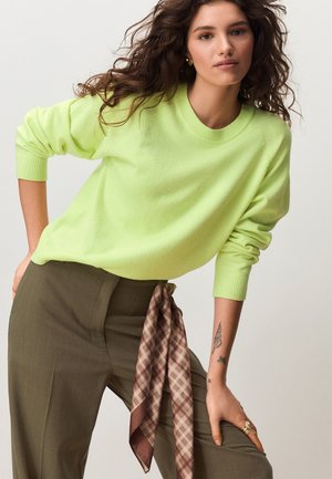 Vrouw met krullend haar draagt een limegroene trui, olijfgroene broek en een bruin geruit sjaaltje dat om haar middel is geknoopt, met tatoeages zichtbaar op haar linkerarm.