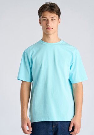 Młody mężczyzna stojący, ubrany w jasnoniebieską koszulkę z krótkim rękawem i ciemne dżinsy na jednolitym jasnoszarym tle.