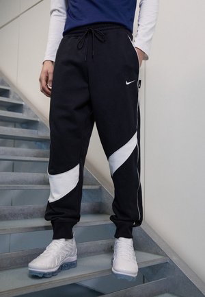 Persona con pantalones jogger Nike en blanco y negro y zapatillas blancas, de pie en una escalera interior con la mano en el bolsillo.