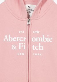 Różowa bluza z zamkiem błyskawicznym, wykonana z miękkiego materiału, z białym napisem "Abercrombie & Fitch EST. 1892 NOWY JORK" wyraźnie widocznym.