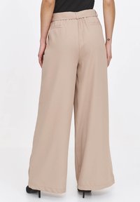 Beżowe spodnie szerokie wykonane z gładkiego materiału. Posiadają wysoki stan, elastyczny pas oraz boczne kieszenie. Ląd zakończenia lekko dotyka ziemi.