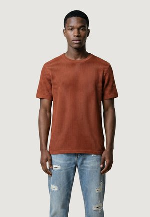 Uomo che indossa una maglietta a maniche corte di colore arancione ruggine con trama e jeans azzurri chiaro strappati, in piedi davanti a uno sfondo chiaro e semplice.