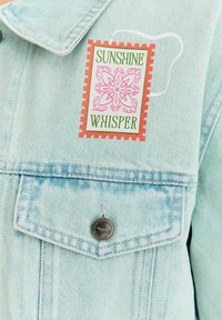 Jasnoniebieska kurtka dżinsowa z naszywką naszytą, przedstawiającą różowy kwiatowy wzór oraz zielony napis "SUNSHINE WHISPER". Widoczne detale z metalowymi guzikami.