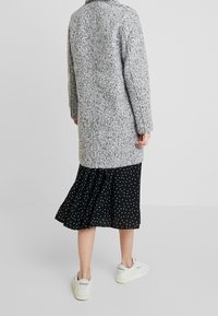 Harmaa, teksturoitu takki suoralla leikkauksella, päällä musta pilkullinen hame ja valkoiset lenkkarit. Neutraali tausta.