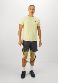 Mann iført gul t-skjorte og svarte shorts som holder en tennisracket, stående mot en enfarget hvit bakgrunn.