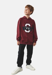 Bordowy hoodie z kapturem z napisem "BRAVE" w kolorze turkusowym, w połączeniu z czarnymi spodniami cargo i białymi sneakerami. Gładki materiał, luźny krój.