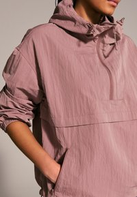 Giacca leggera rosa chiaro realizzata in tessuto testurizzato, con colletto a zip, cappuccio regolabile e tasche laterali. Polsini e orlo elastici.