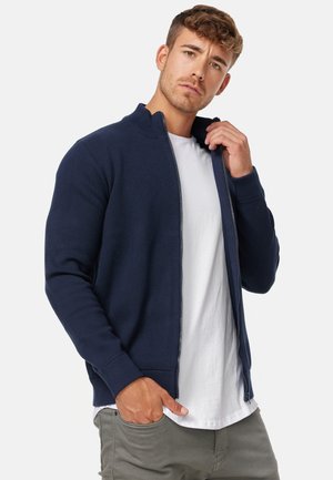 Navy zip-up sweatshirt with a high collar, ribbed cuffs, and side pockets, worn over a white t-shirt. Soft fabric with a fitted design.