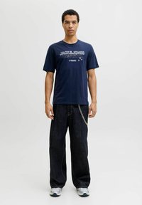Camiseta de algodón azul marino con texto gráfico blanco, combinada con pantalones de denim oscuros de pierna ancha que presentan un detalle de cadena en los bolsillos delanteros.
