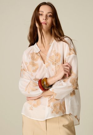 Jonge vrouw met lang bruin haar die een witte blouse draagt met beige bloemmotief en twee dikke armbanden om haar pols.