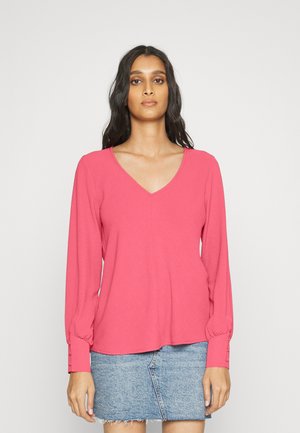 Blusa de manga longa, de gola em V, em rosa brilhante, feita de material macio. Apresenta detalhes em botão nos punhos e um corte descontraído. Combinada com uma saia de ganga.