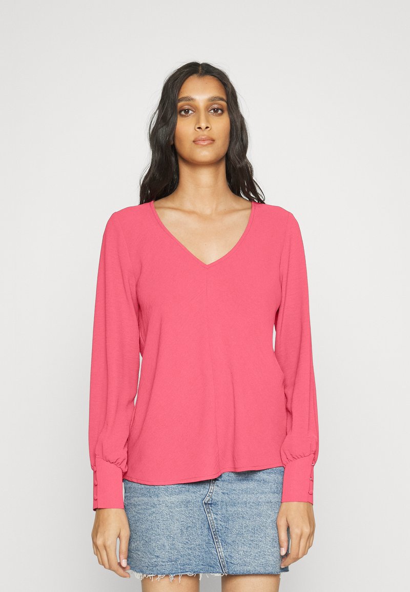 Blusa de manga longa, de gola em V, em rosa brilhante, feita de material macio. Apresenta detalhes em botão nos punhos e um corte descontraído. Combinada com uma saia de ganga.