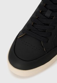 Black leather sneaker with textured surface, round toe, and perforations. Features black laces and a rubber tan sole with a patterned edge.