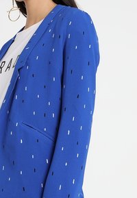 Královsky modrý blazer vyrobený z měkké látky s rozptýlenými černobílými tečkami, s klasickým klopou a jednou přední kapsou.