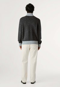 Maglione grigio a maglia grossa con colletto alto e polsini a coste, abbinato a pantaloni color crema e scarpe nere, visto da dietro.