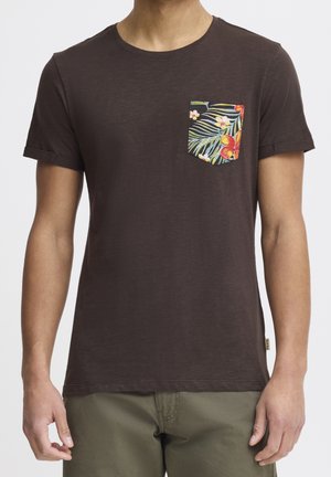 Mann trägt ein dunkelbraunes kurzärmliges T-Shirt mit einer bunten tropischen Blumentasche auf der linken Brust, kombiniert mit olivgrüner Hose.