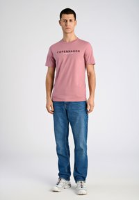 Ljusrosa T-shirt med texten "COPENHAGEN", kombinerad med blå jeans och vita sneakers. T-shirten har korta ärmar och en rund halsdesign.