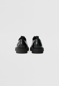 Zapatos de cuero negro con una textura lisa y una suela de goma gruesa. Incluyen una lengüeta en la parte trasera y sutiles detalles de costura.