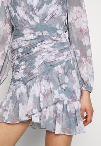 Květinové mini šaty z měkké texturované tkaniny. Světle modrozelený základ s bílými a fialovými květy. Má nařasený, plisovaný design a volánkový lem.