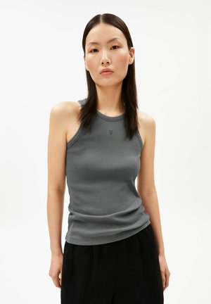 Femme aux cheveux raides et foncés portant un haut gris côtelé sans manches et un pantalon noir, debout devant un fond blanc uni.