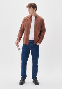 Camicia marrone a maniche lunghe con bottoni, t-shirt bianca sotto, jeans di denim blu, occhiali da sole in mano, scarpe nere, in piedi contro uno sfondo bianco.