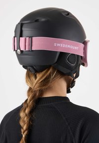 Musta hiihtokypärä, jossa on vaaleanpunainen hihna, teksturoitu mattapinta, ilmanvaihtoaukot ja säädettävä solki. Takakuvassa näkyy lettiin punottu poninhäntä.