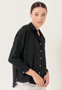 Camicia nera in lino con maniche lunghe, chiusura con bottoni sulla parte anteriore e colletto. Presenta bottoni chiari e una fessura laterale per facilitare il movimento.