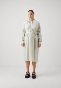 Robe rayée vert clair avec un col, des manches longues, une taille cintrée par un lien, et de longueur mi-mollet. Associée à des sandales noires épaisses.