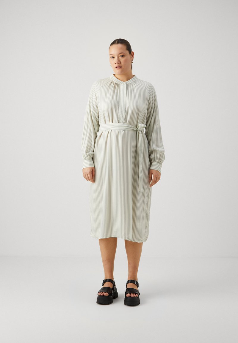 Rochie verde deschis cu dungi, cu guler, mâneci lungi, talie strânsă cu o fundă, și lungime până la jumătatea gambei. Asortată cu sandale negre cu talpă groasă.