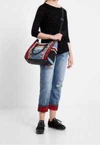 Person som bär en svart topp, uppvikta blå jeans med röd rutigt foder, svarta sneakers och bär en stor patchwork-väska med denim- och rutmönster.