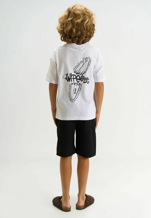Göndör szőke hajú gyermek fehér "Wipeout" feliratú pólóban, fekete rövidnadrágban és barna szandálban, fehér háttér előtt állva.