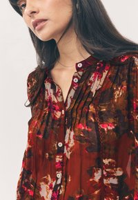 Blusa floreale marrone con pieghe, scollo rotondo e bottoni bianchi. Il tessuto ha una consistenza morbida e trasparente con motivi vivaci rosa e verdi.