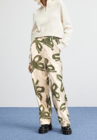 Person som bär en krämfärgad halvdraget tröja, beigefärgade kordjeans med grön abstrakt blommönster, och svarta klumpiga plattformsandaler på en blå matta.