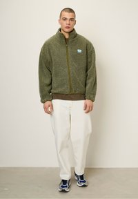 Chaqueta de forro polar verde oliva con cremallera frontal, puños elásticos y un pequeño logo en parche. Combinada con una capa interior marrón y pantalones blancos.