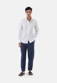Weißes Langarm-Hemd mit dezenten Streifen, kombiniert mit navyblauen Leinenhosen und schwarzen flachen Sandalen.