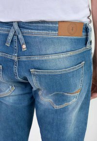Jeans de mezclilla en azul claro con un acabado desgastado. Presenta un parche de cuero en la parte trasera de la cintura y dos bolsillos traseros con costuras curvas.