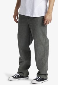 Hombre que lleva pantalones de pana gris holgados, zapatillas negras y blancas, y una camiseta blanca, de pie con las manos relajadas a los lados.