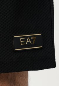 Μαύρο υφασμάτινο ύφασμα με ανάγλυφη υφή παρουσιάζει μια εμφανή ταμπέλα με το λογότυπο EA7 Emporio Armani σε μαύρο και χρυσό, με μοντέρνο σχέδιο και καθαρή γραμματοσειρά.