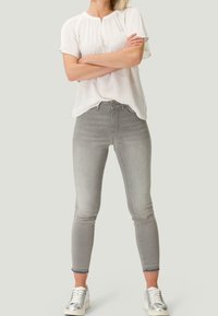 Světlé šedé slim-fit džíny s vysokým pasem, vybavené knoflíkovým uzávěrem a jemným vyblednutím. V kombinaci s bílou blouse s krátkým rukávem a stříbrnými teniskami.