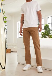 Weißes Baumwoll-T-Shirt, beige Chinos und weiße Sneaker. Die Umgebung ist hell mit großen Fenstern und minimalistischem Mobiliar.