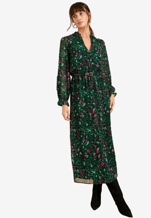 Green floral print maxi dress with long, sheer sleeves and a belted waist. Features a v-neckline and flowing silhouette. Black knee-high boots.