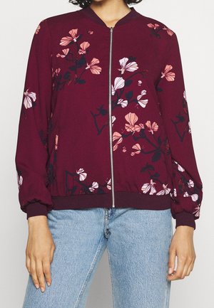 Frau trägt eine weinrote Blumen-Zipperjacke mit pinken und weißen Blumen und hellblauen Jeans.