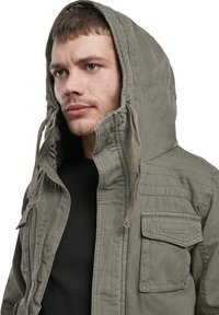 Grüne Kapuzenjacke mit einem Reißverschluss vorne, strukturiertem Stoff, großen Fronttaschen und Kordeln an der Kapuze. Über einem schwarzen Shirt getragen.