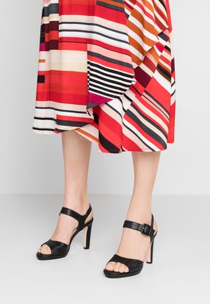 Sorte højhælede sandaler med ankelrem, der har en tekstureret overdel og åben tå-design, bæres med en flerfarvet, let kjole med striber.