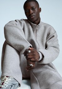 Giovane uomo con pelle scura, vestito con tuta leggera grigia e sneakers, seduto con le mani unite e che guarda in avanti su uno sfondo semplice.