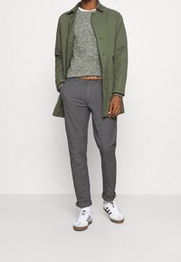 Grüner leichter Mantel über einem strukturierten grauen Pullover, graue Hose und weiße Sneakers mit schwarzen Streifen, dazu ein brauner Gürtel.