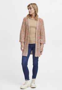 Randig kofta i jordnära färger av orange, beige och kräm, med en lös passform, långa ärmar, ribbade mudd och en öppen frontdesign.