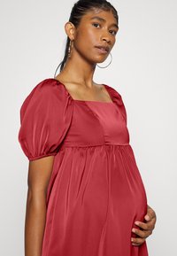 Vestido de maternidade em cetim vermelho com mangas bufantes, decote quadrado e cintura franzida. A textura suave e o design fluido destacam o conforto.