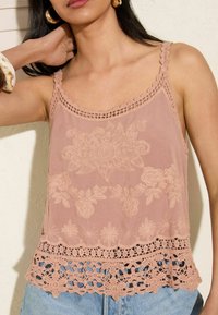 Top senza maniche di un rosa chiaro, con motivi floreali ricamati, bordo in pizzo e orlo sagomato. Il tessuto sembra leggero e delicato.