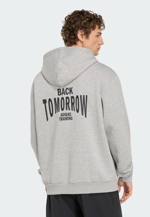 Junger Mann mit lockigem Haar, der einen hellgrauen Adidas-Hoodie mit dem Aufdruck „BACK TOMORROW ADIDAS TRAINING“ auf dem Rücken trägt, seitlich blickend.
